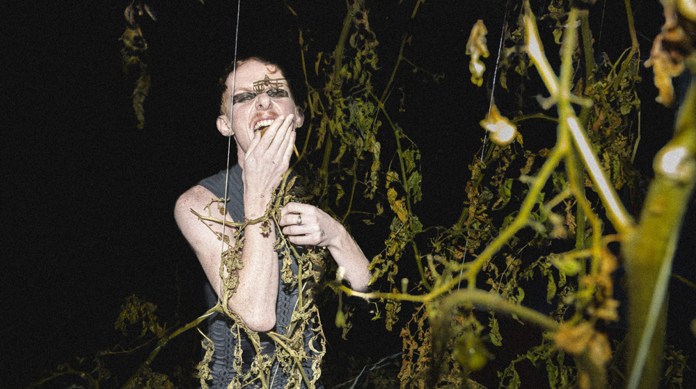 A person stands in the darkness among vegetation, with a hand in front of their mouth.