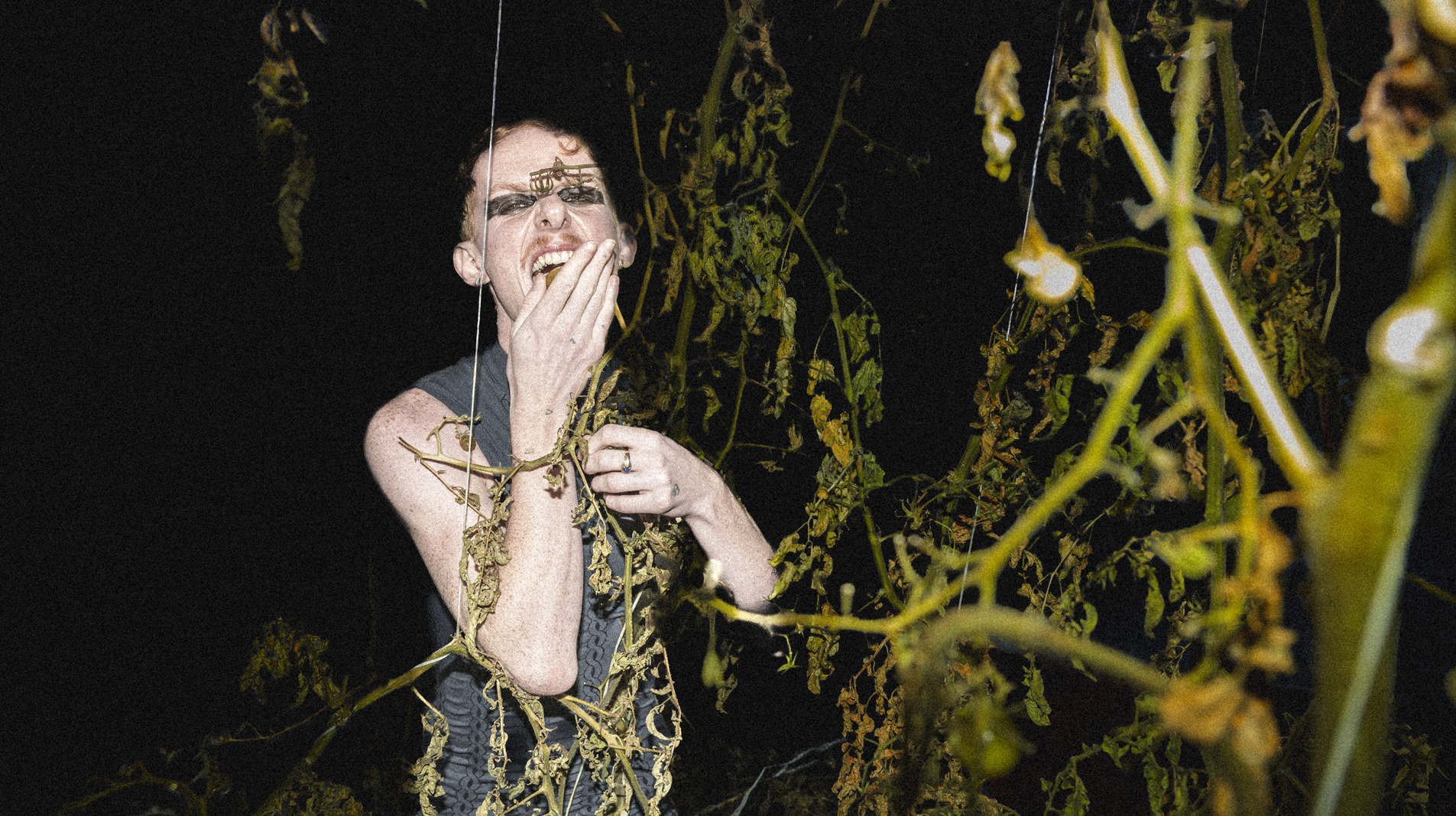 A person stands in the darkness among vegetation, with a hand in front of their mouth.