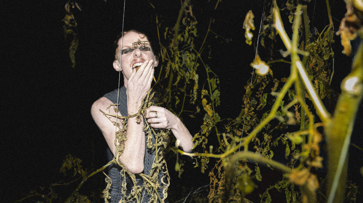 A person stands in the darkness among vegetation, with a hand in front of their mouth.