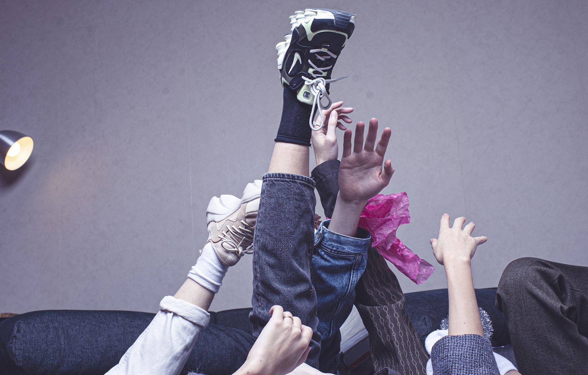Several hands and legs from different people reach upward in front of a gray wall, with parts of a sofa and a lamp visible in the background.